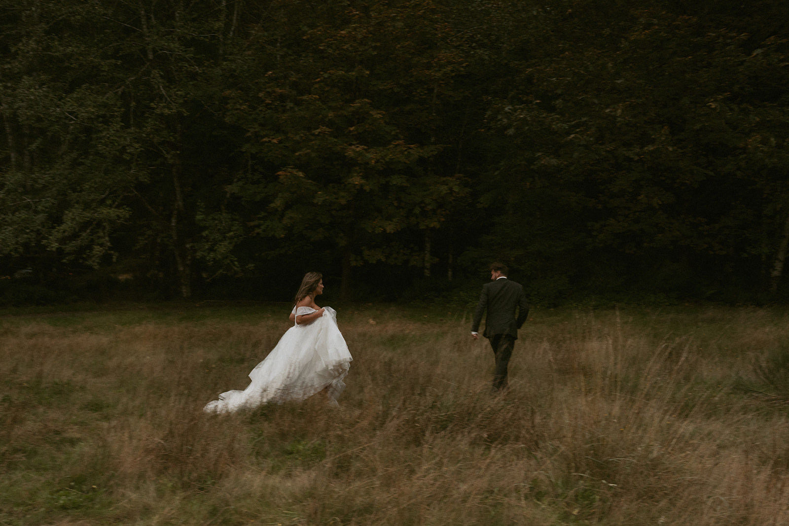 The couple walks through a golden meadow at dusk, the bride holding up her gown as they wander toward the trees.