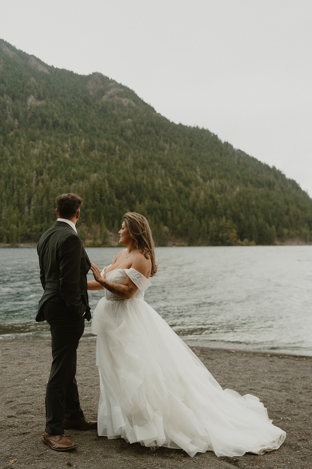 A couple stands close together, holding hands beside a misty lake with lush, green mountains in the background.