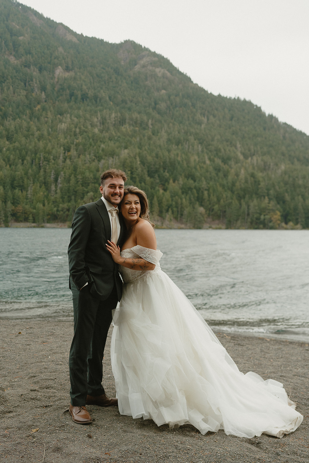 The couple stands together in joyful embrace by a peaceful mountain lake, surrounded by evergreen forests.