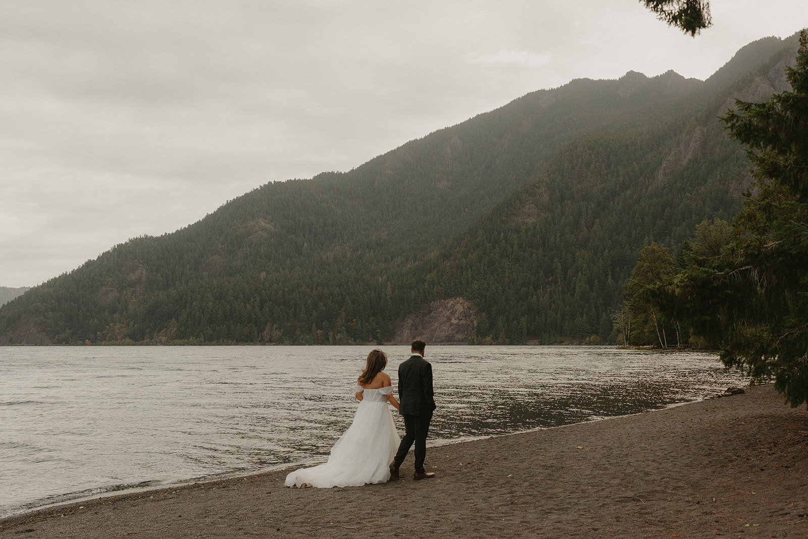A bride and groom walk along the shoreline of a peaceful lake, surrounded by forested mountains under an overcast sky during their Olympic National Park elopement.
