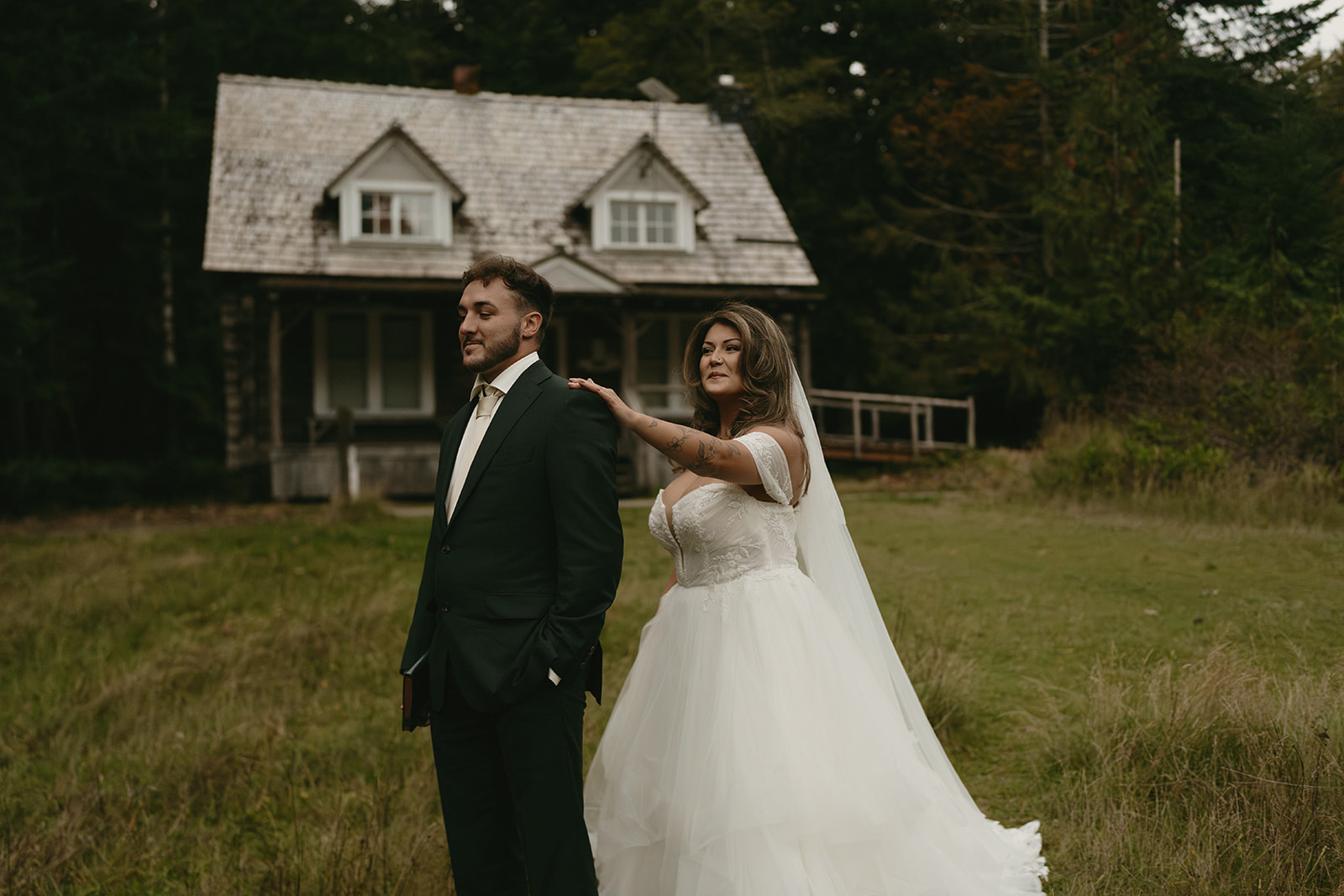 The bride reaches out to tap the groom on the shoulder during their first look outside a rustic cabin in the woods.