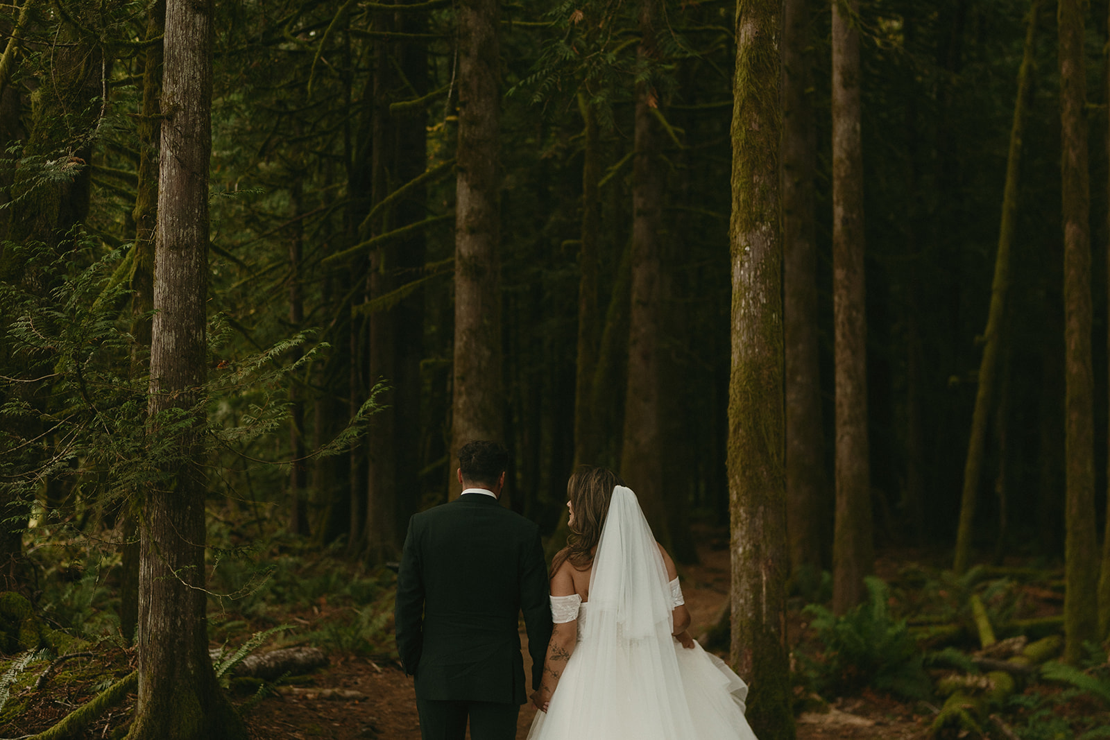 The couple walks hand-in-hand into the dense forest, their wedding attire standing out against the moss-covered trees during their Olympic National Park elopement.
