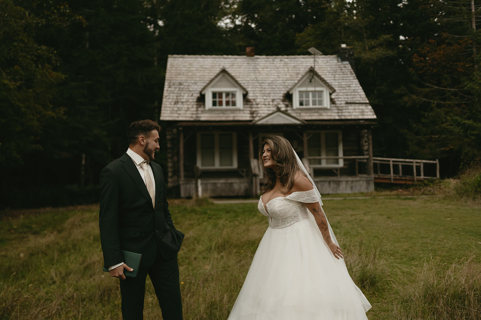 The couple smiles warmly at each other in front of a weathered forest cabin, moments after their first look during their Olympic National Park elopement.