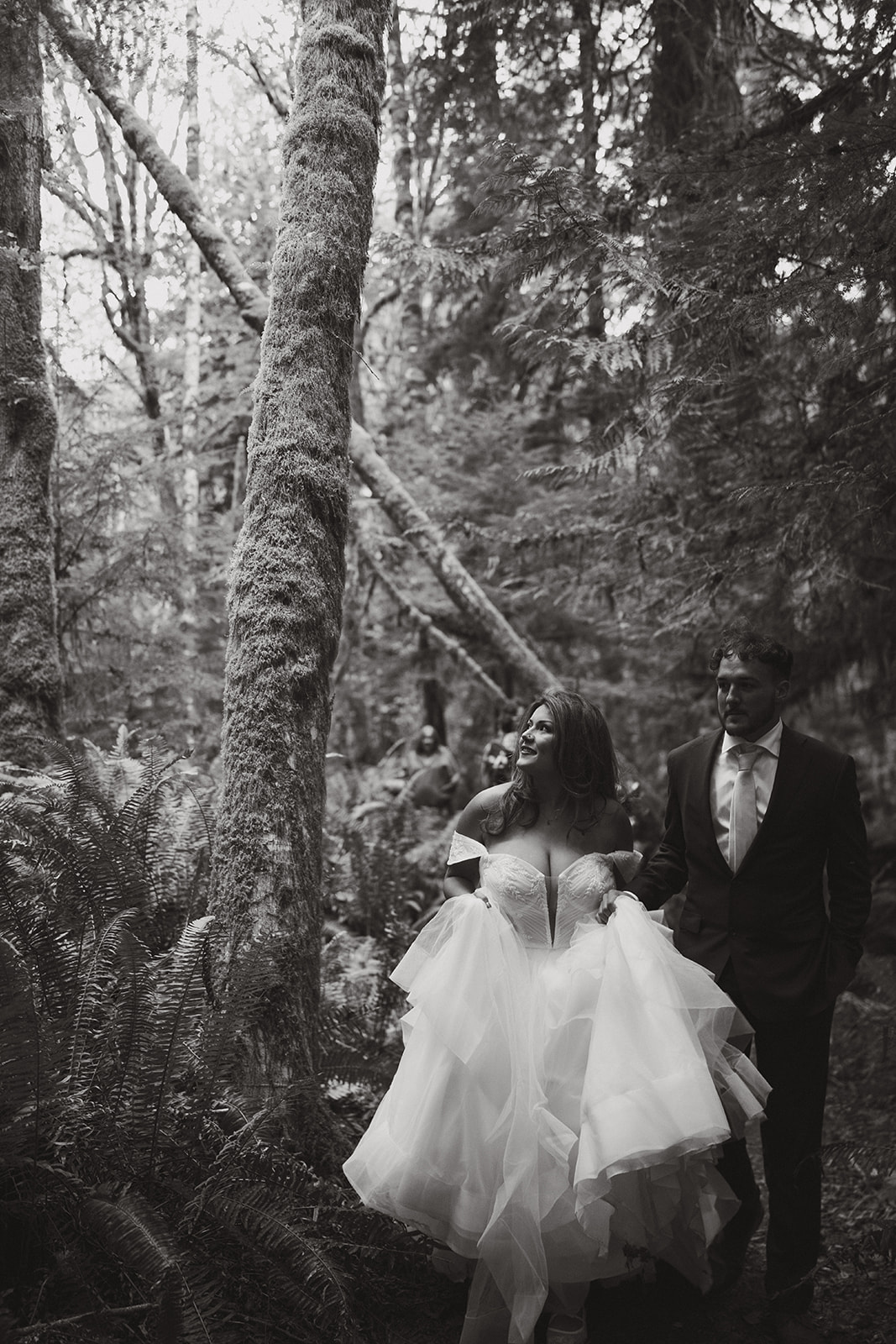 A black and white image of the bride lifting her gown as she and the groom walk through a lush, forested trail, light filtering through the trees.
