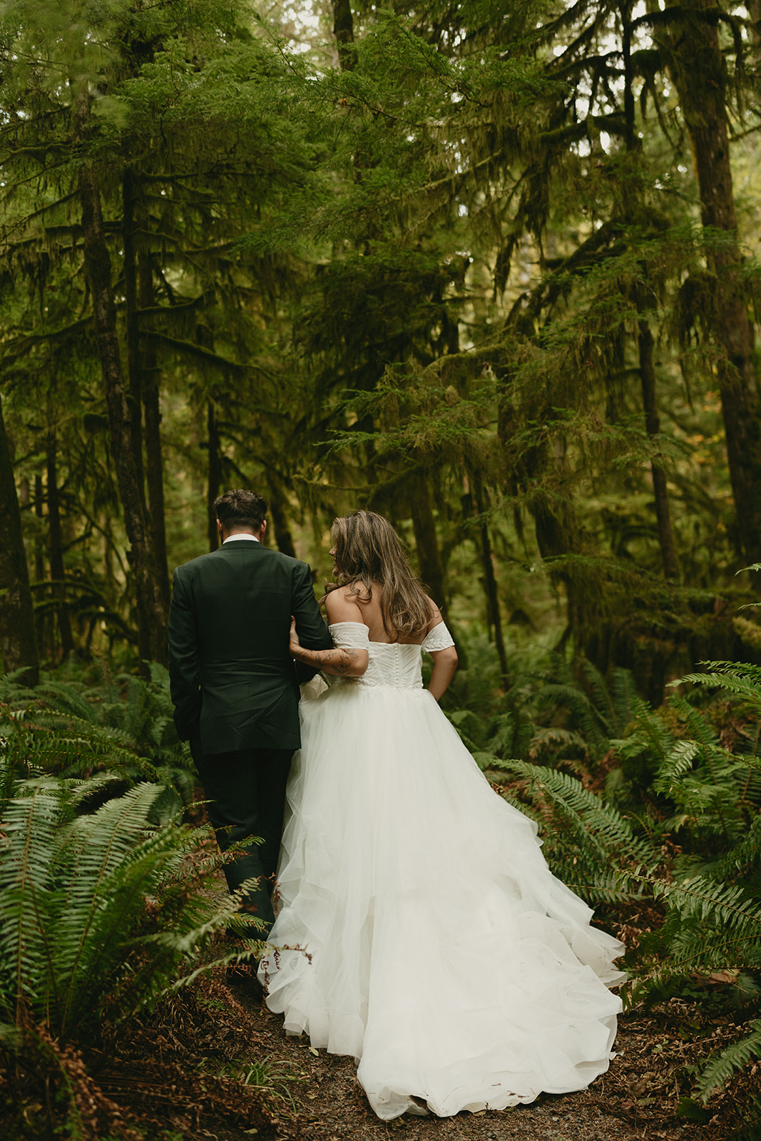 The bride lifts her gown slightly as she and the groom stroll a shaded woodland path, lost in the moment together.