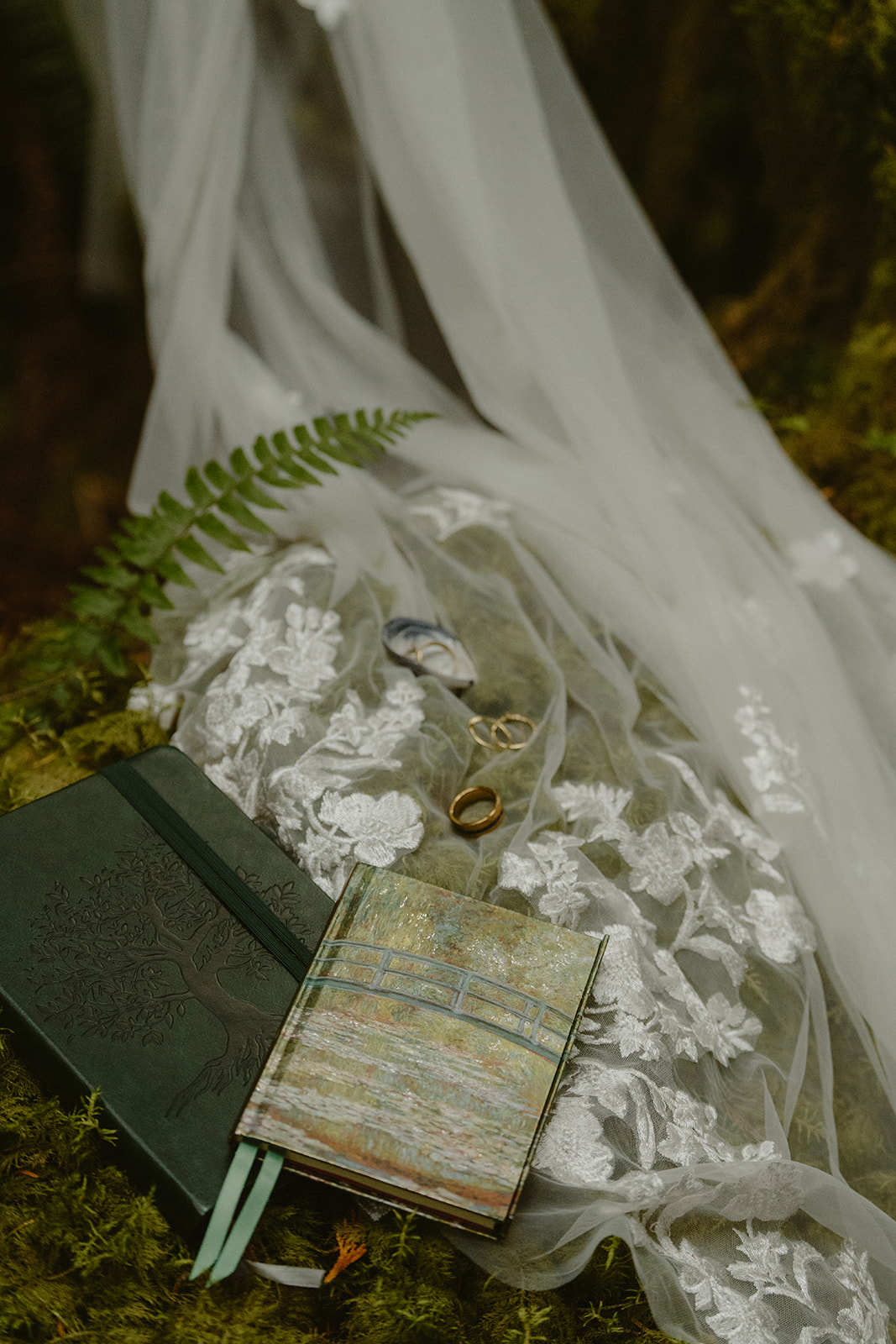Flat lay of vow books, wedding rings, and a lace veil carefully placed on a mossy forest floor, capturing the quiet details of an elopement day.