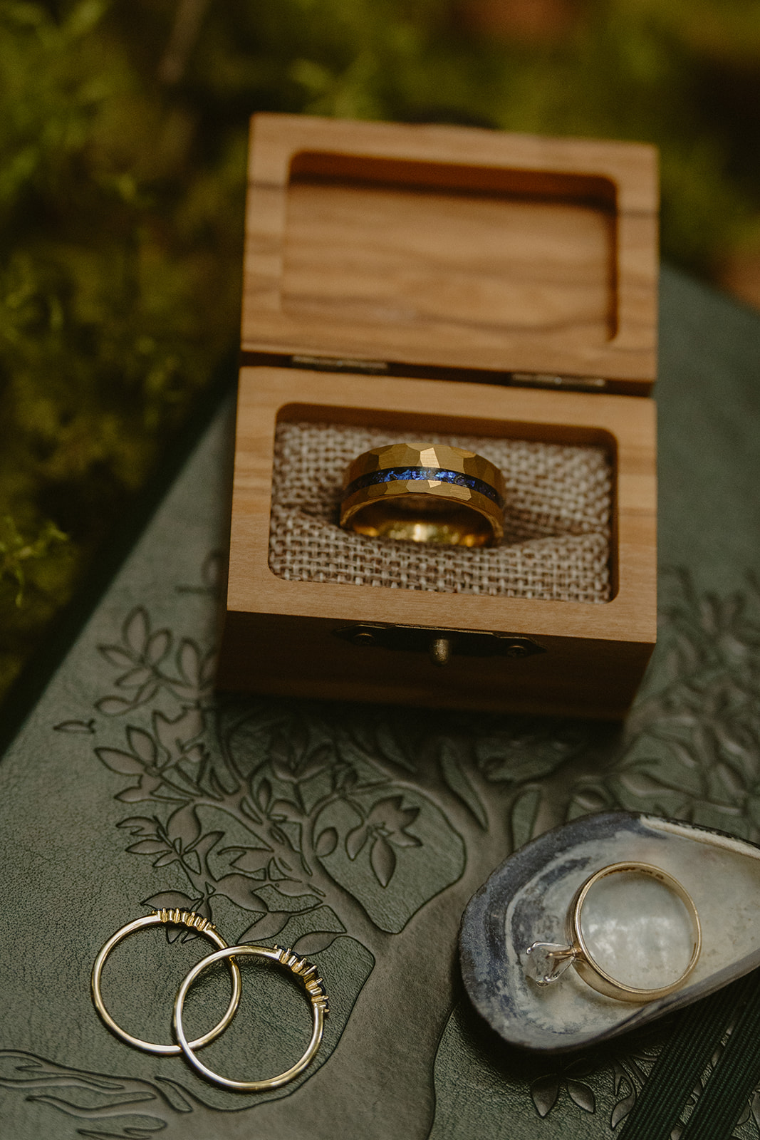 A wooden ring box and wedding rings rest on a forest-green vow book, styled thoughtfully in the mossy woods during an Olympic National Park elopement.