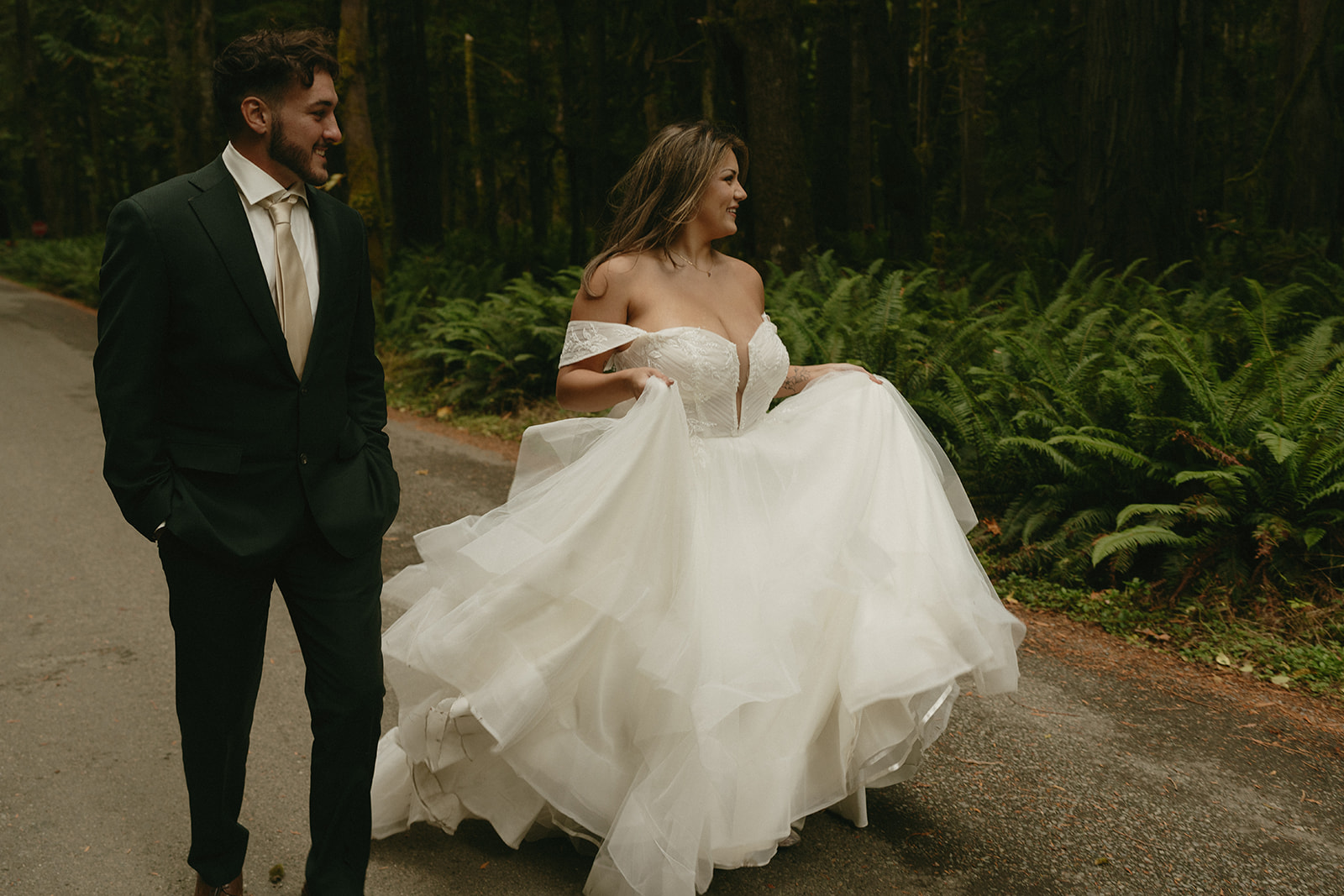 The couple walks side by side down a forest road, the bride’s gown flowing around her as they smile together during their Olympic National Park elopement.