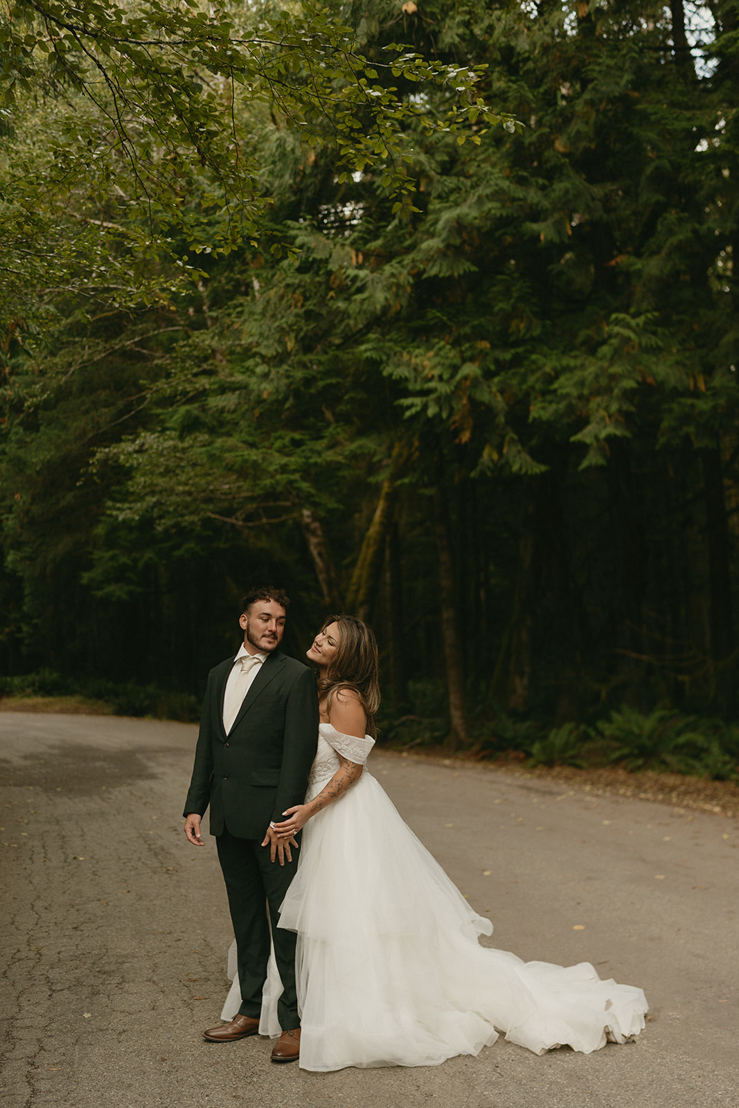 The groom gently adjusts the bride’s dress as they pause on a tree-lined road, surrounded by towering evergreens during their Olympic National Park elopement.