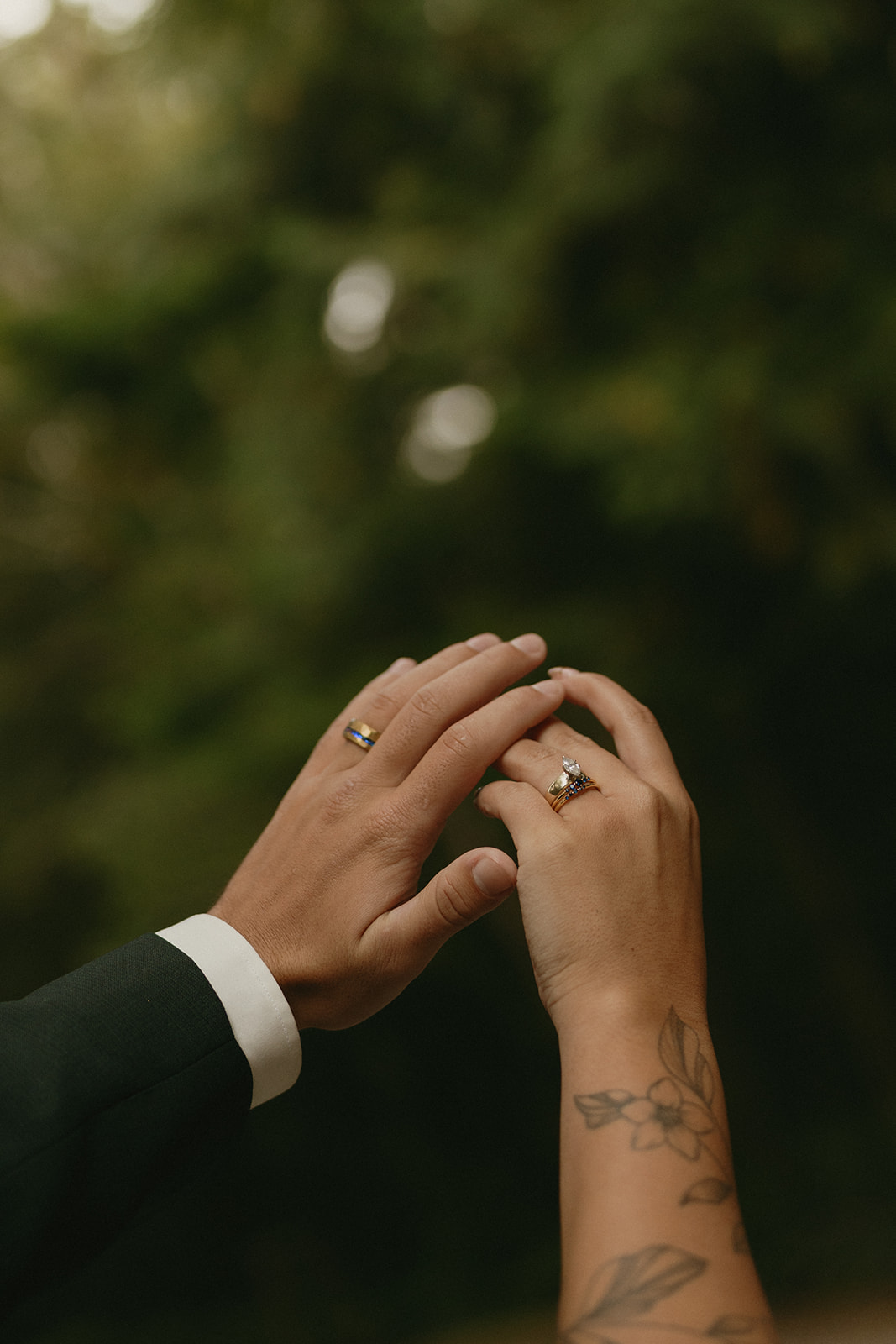 Hands raised together, the couple shows off their wedding rings with the forest canopy blurred softly in the background.