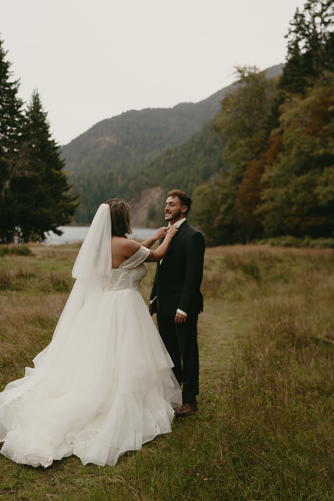 The bride adjusts the groom’s boutonniere with a loving smile, the mountains rising softly behind them.