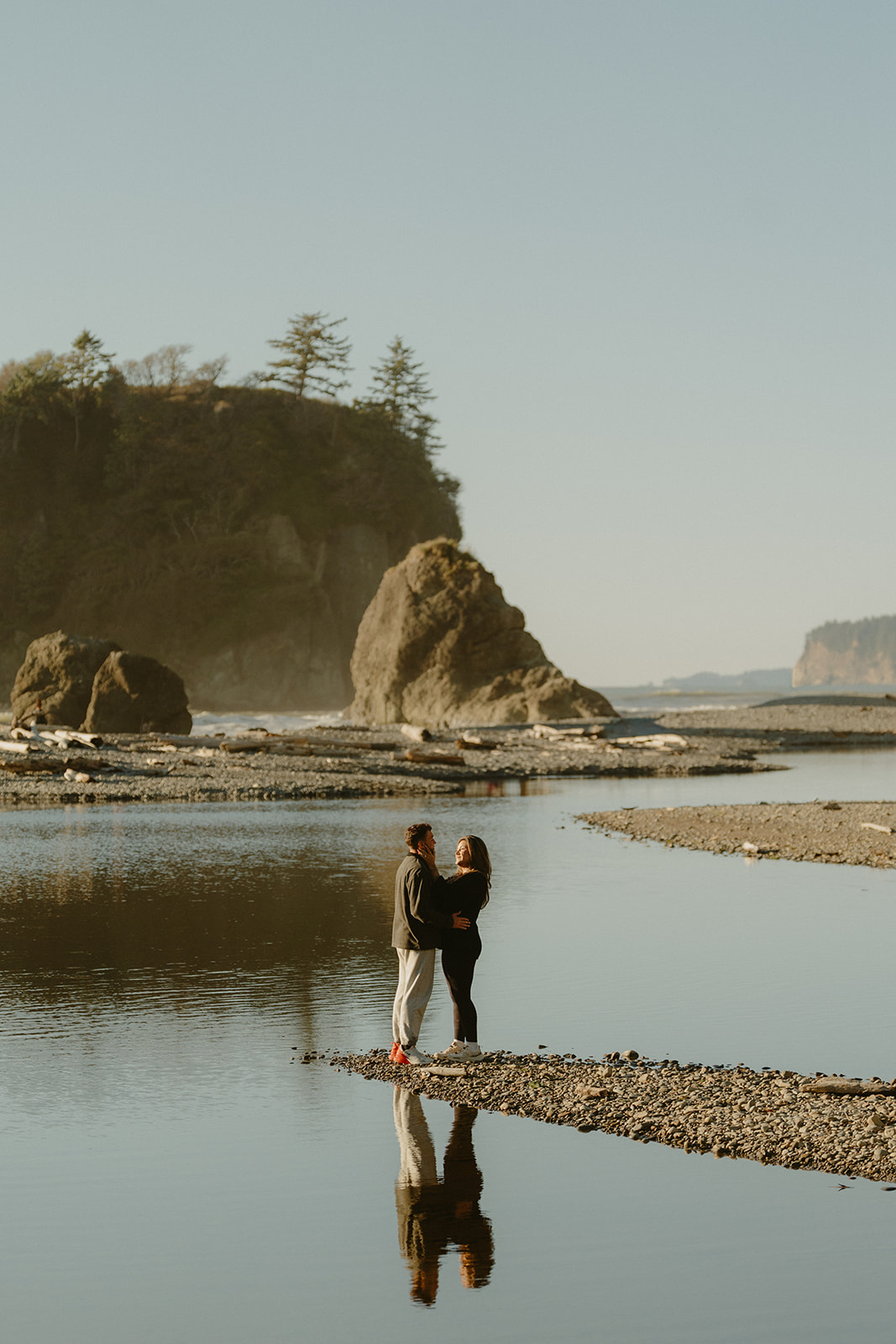 A couple stands together in the stillness of a tidepool, surrounded by sea stacks and soft golden light on the Washington coast.