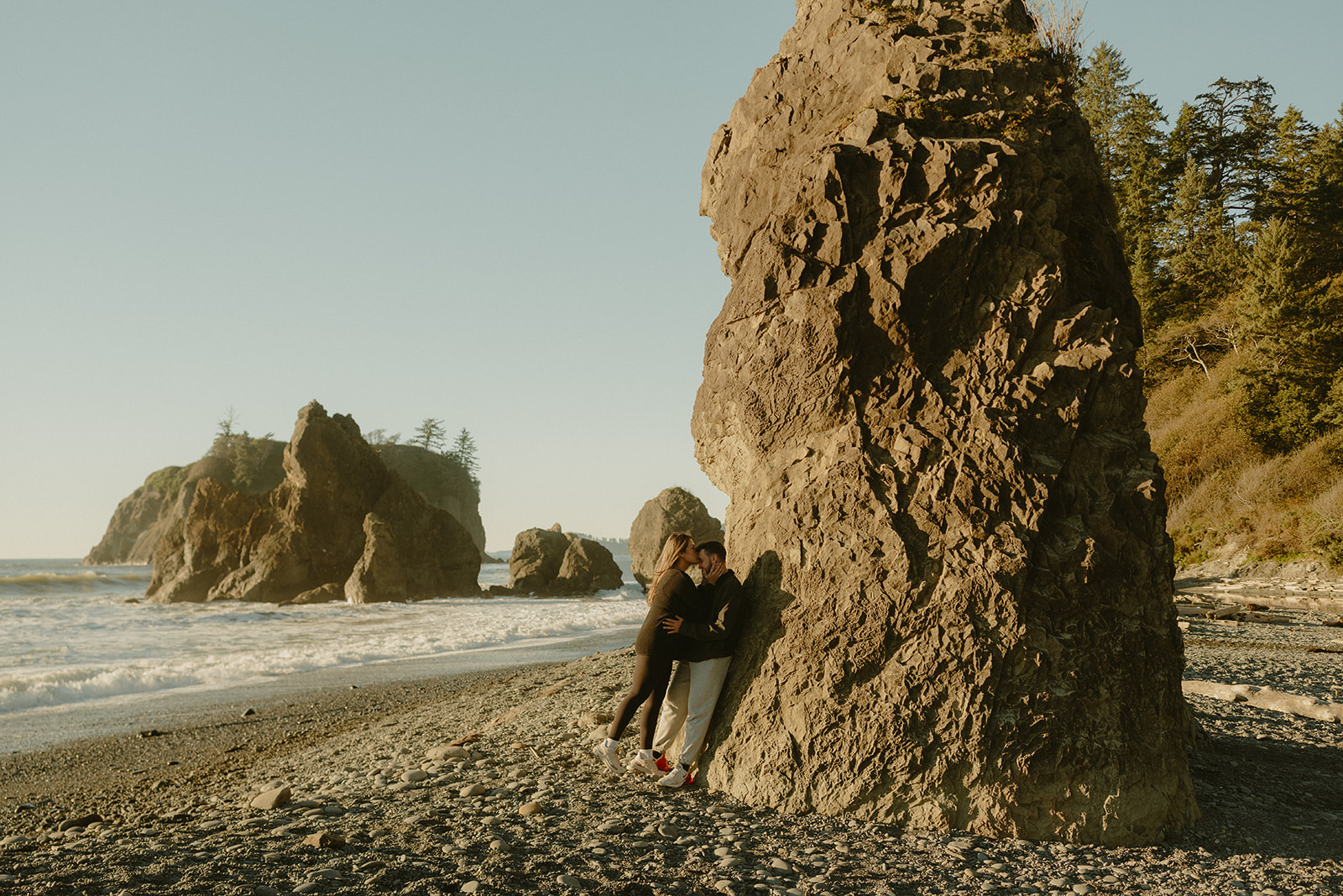 The couple shares a kiss while standing beside a towering rock formation on a rocky shoreline during their coastal Olympic National Park elopement.