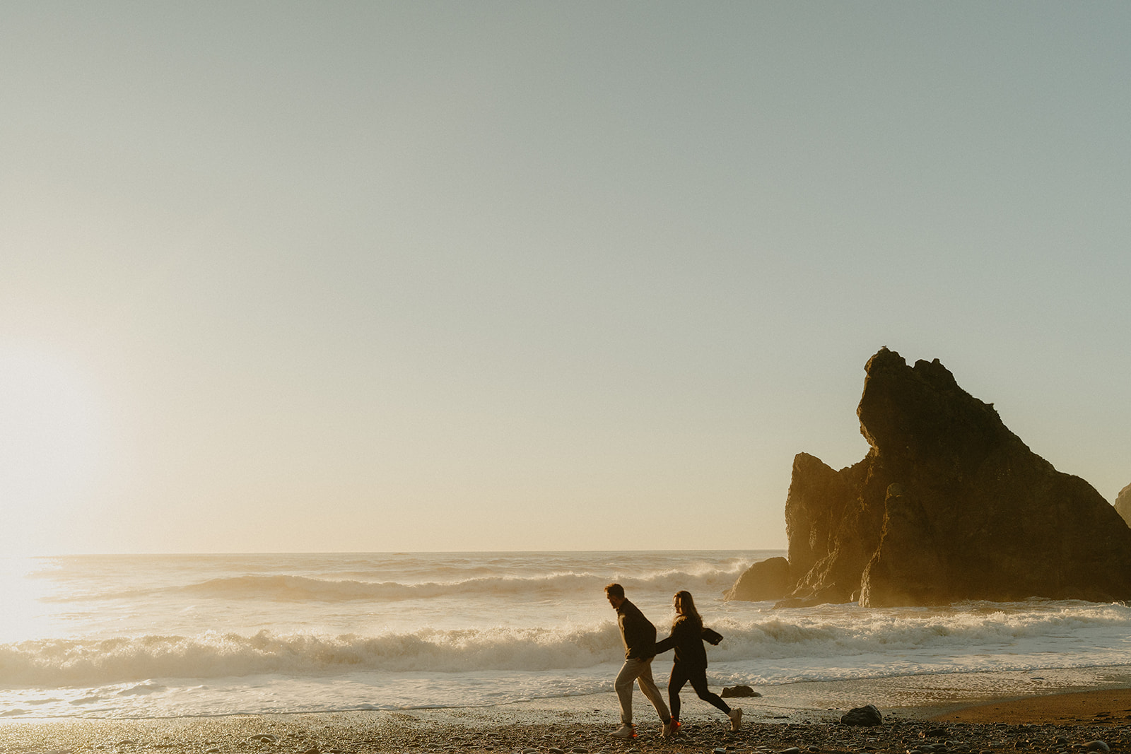 Two people run hand-in-hand along the edge of the ocean at sunset, waves crashing behind them and sea stacks rising in the distance.