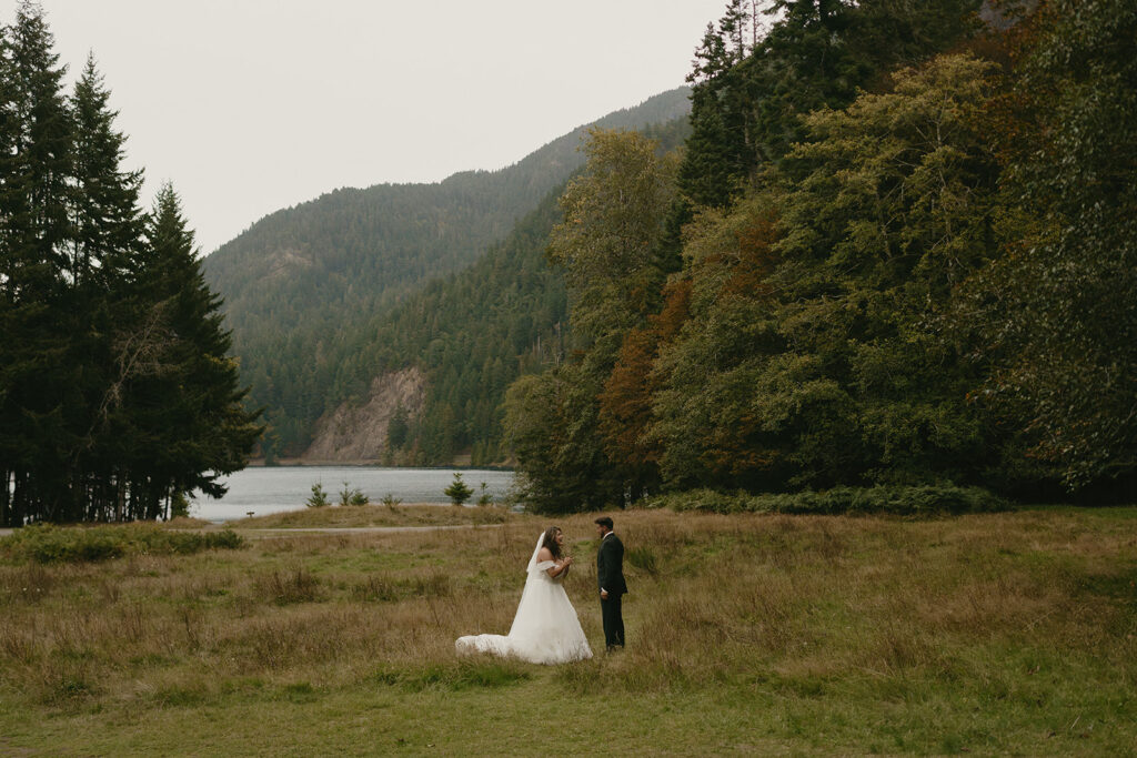 The couple stands in a wide, grassy meadow facing each other with a forested lake and mountains in the distance—an intimate Olympic National Park elopement moment.