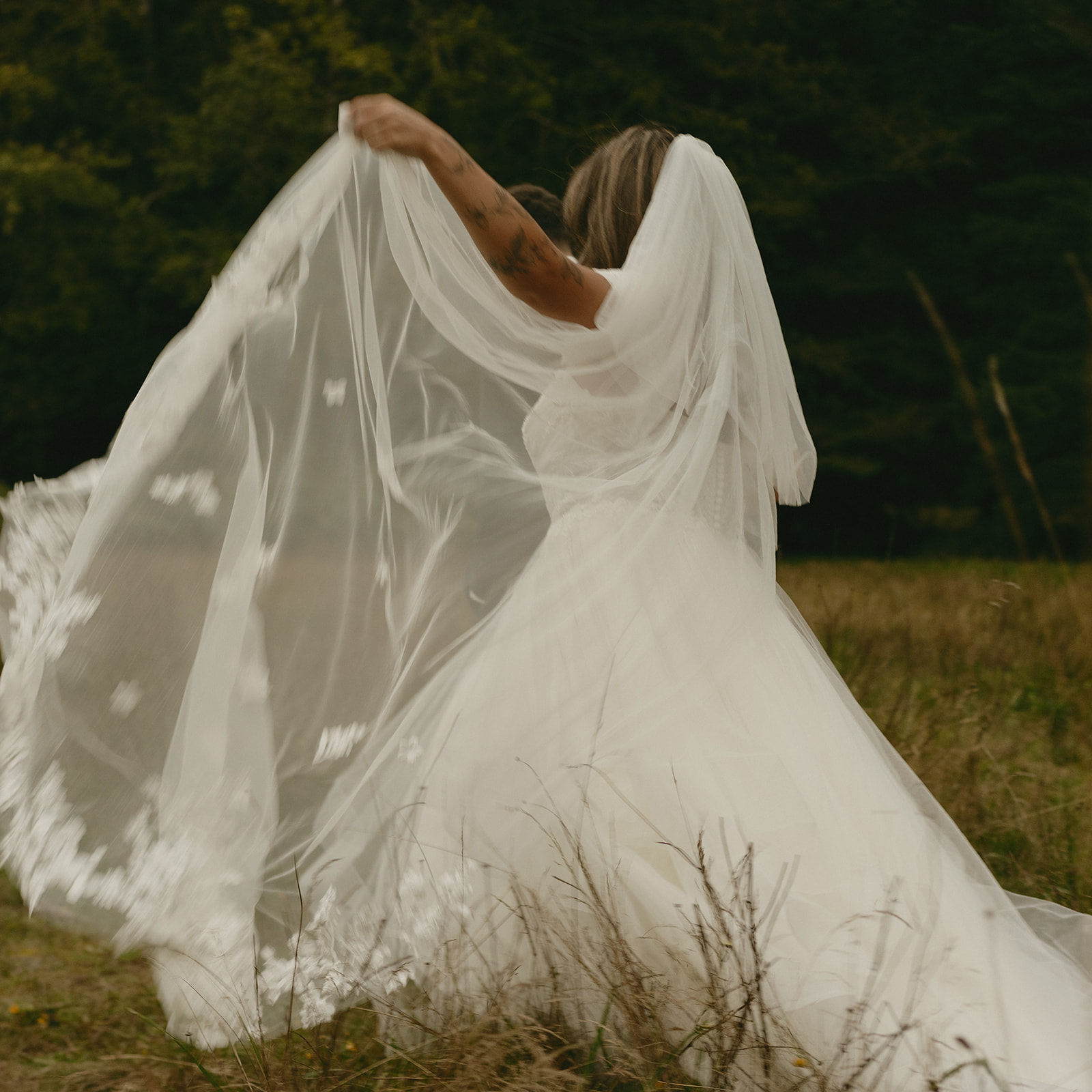 A soft moment as the bride lifts her veil, the fabric catching the wind in a meadow surrounded by quiet greenery.