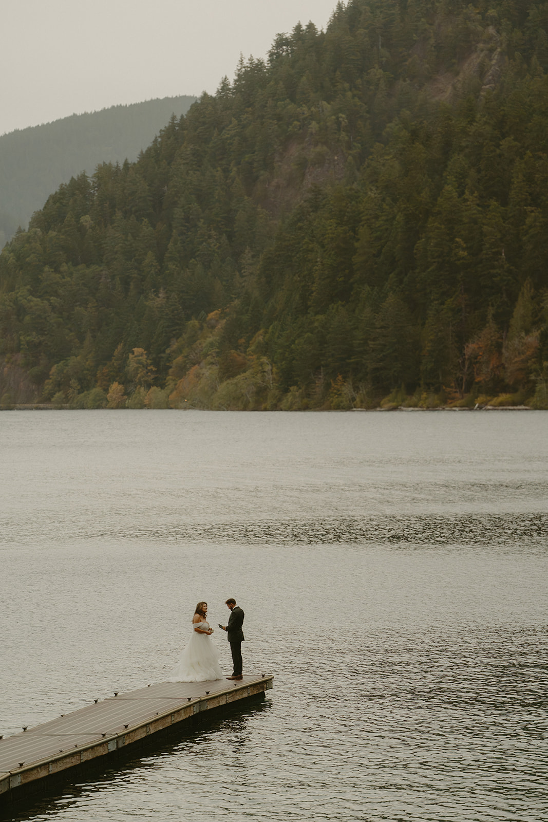 The couple stands on a dock over a quiet lake, surrounded by forested hills in the Pacific Northwest.