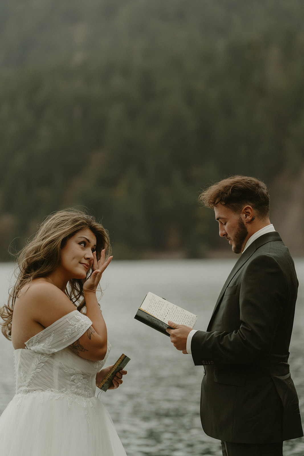 The groom reads vows as the bride wipes away tears, standing beside a lake during their emotional Olympic National Park elopement.