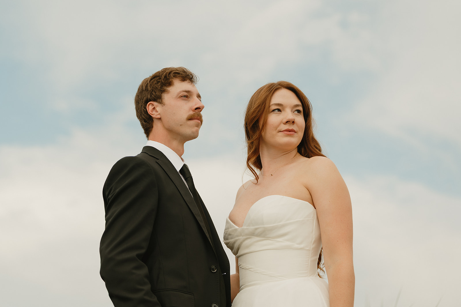 Bride and groom stand side by side, gazing softly into the distance under a pale Washington sky.