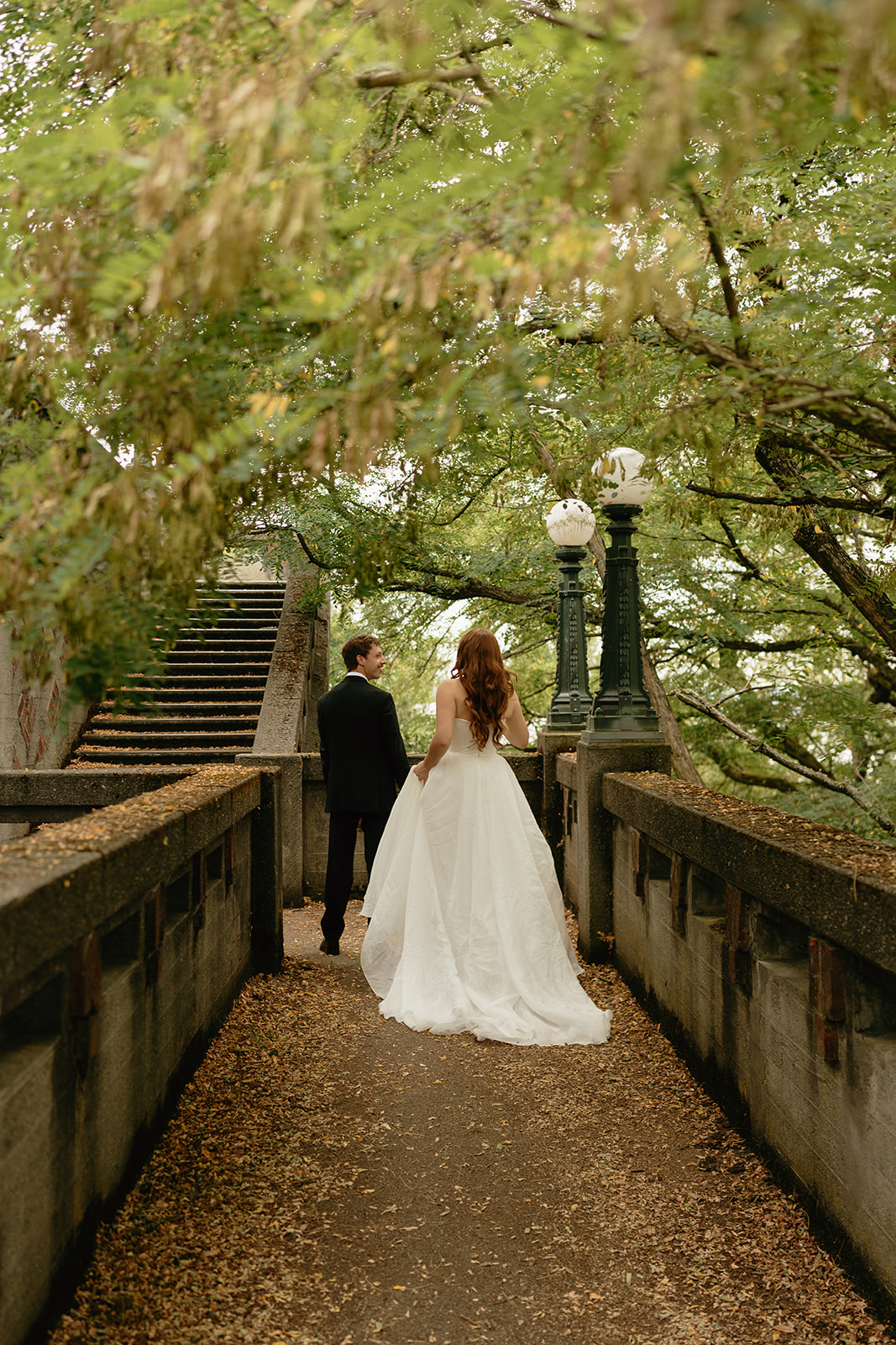 Bride and groom walking hand in hand down a leaf-covered path beneath the trees, surrounded by stillness.