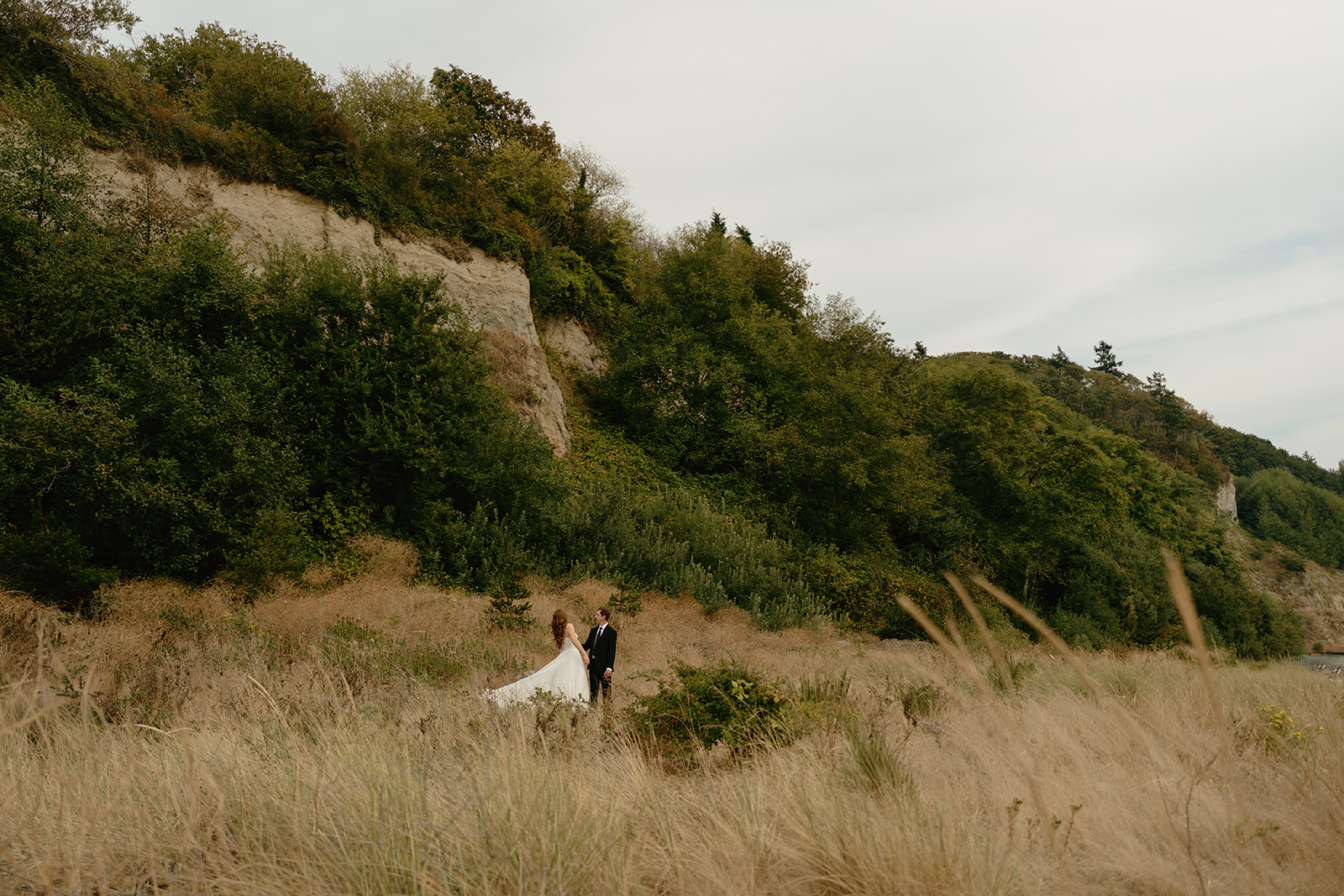 Bride and groom walking hand in hand through tall golden grass beneath rugged coastal cliffs during their intimate wedding in Washington.