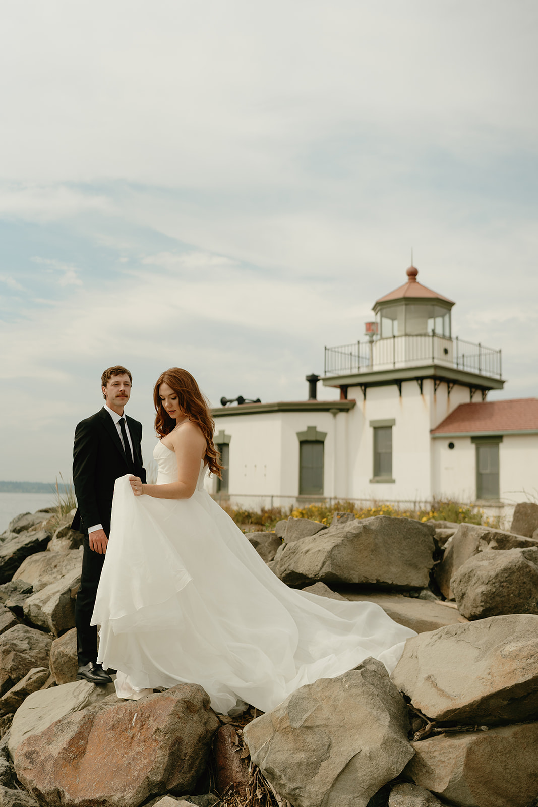 Romantic portrait of a couple standing on coastal rocks with a historic lighthouse in the background during their wedding in Washington.