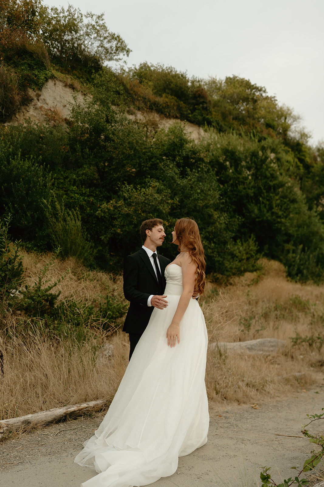 Intimate portrait of a couple standing near the base of a Washington cliffside, surrounded by coastal grasses.