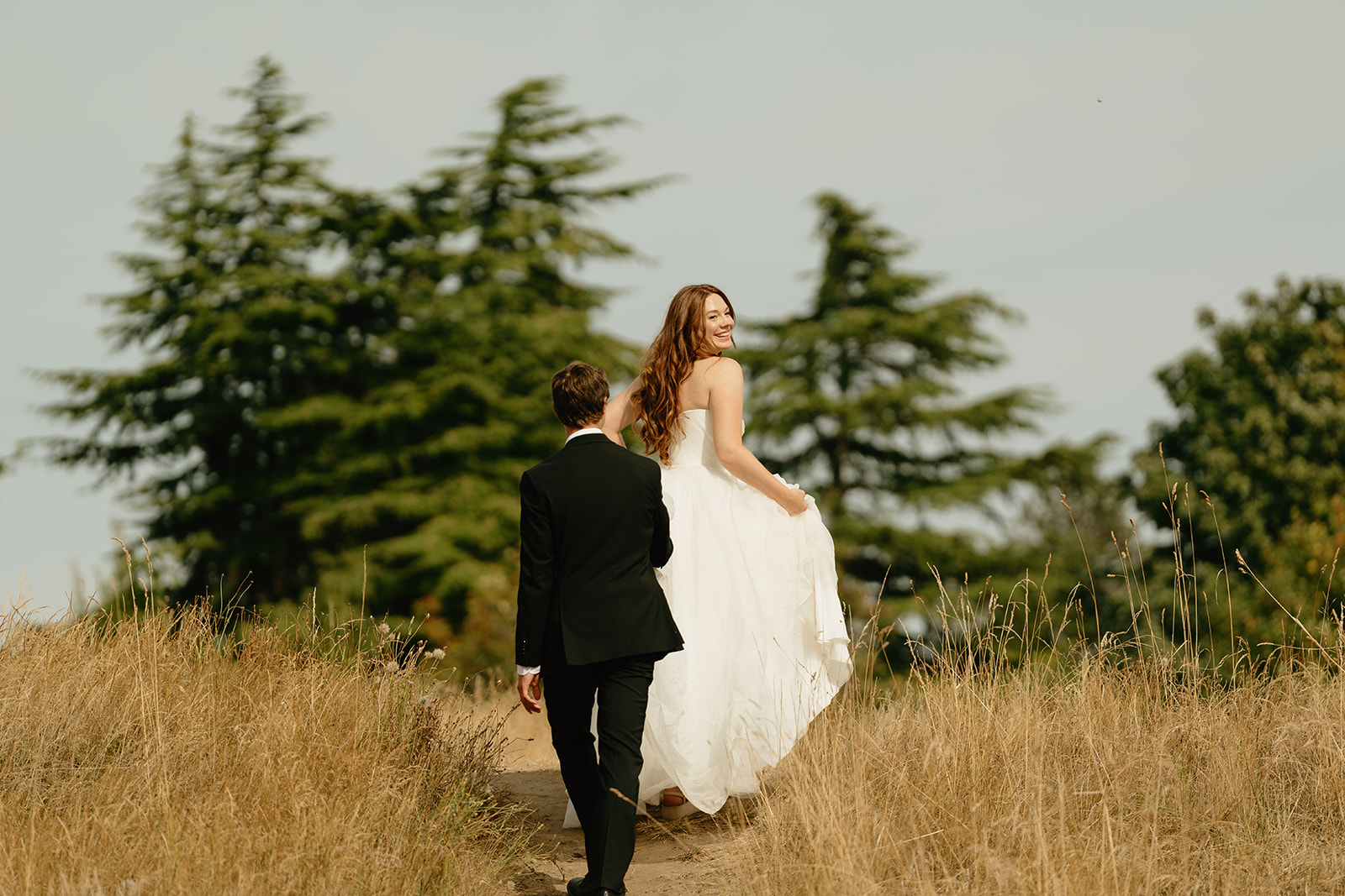 Bride looks over her shoulder with a joyful smile as she walks a sunlit trail with her groom during their wedding in Washington.
