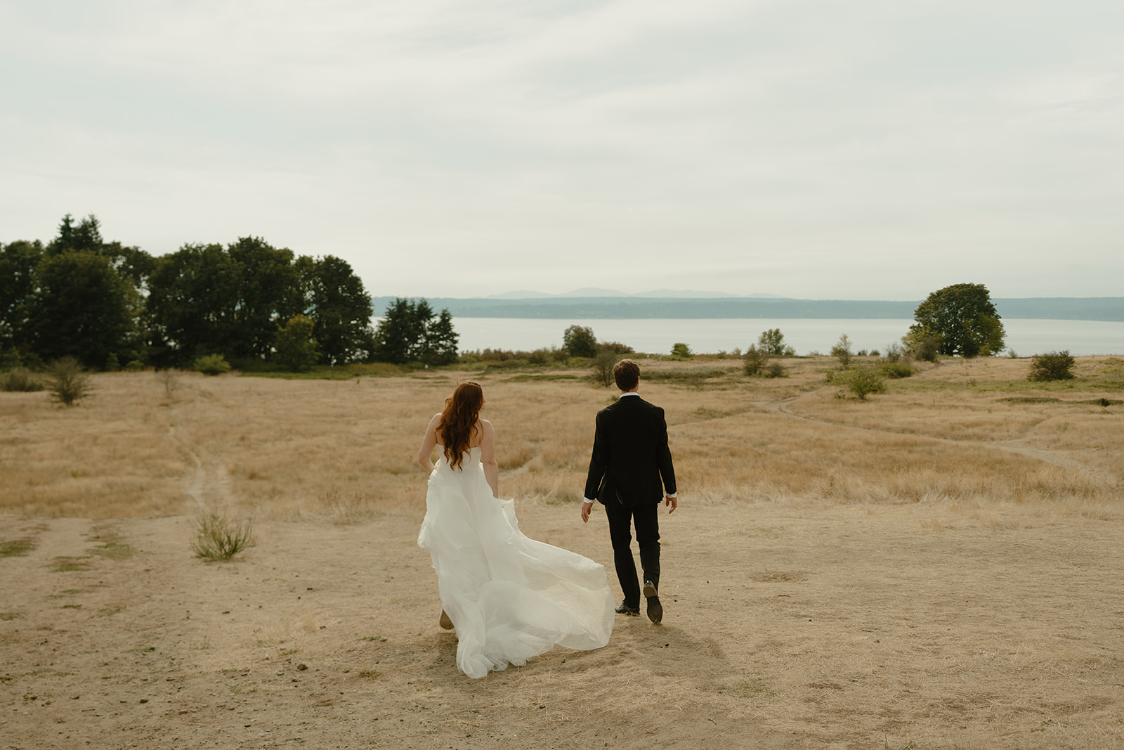 Bride and groom walking across an open golden field toward the water, surrounded by the calm and beauty of their wedding in Washington.