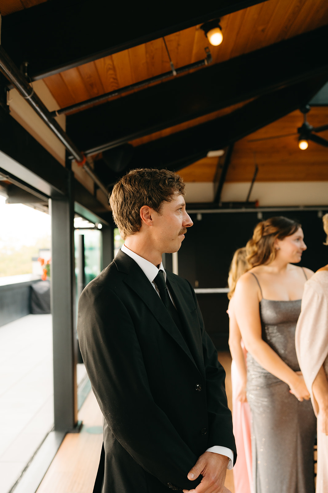Groom waits quietly at the altar, standing beneath warm wooden beams and soft lighting as guests arrive.