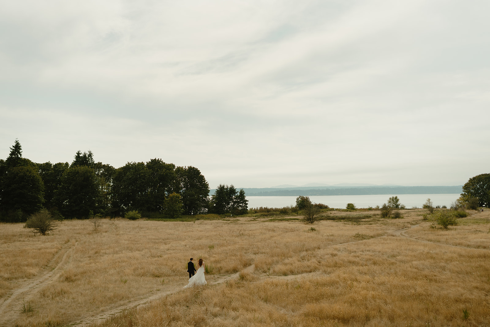 Wide shot of a couple walking through an open meadow near the water, wrapped in the calm of their wedding day.