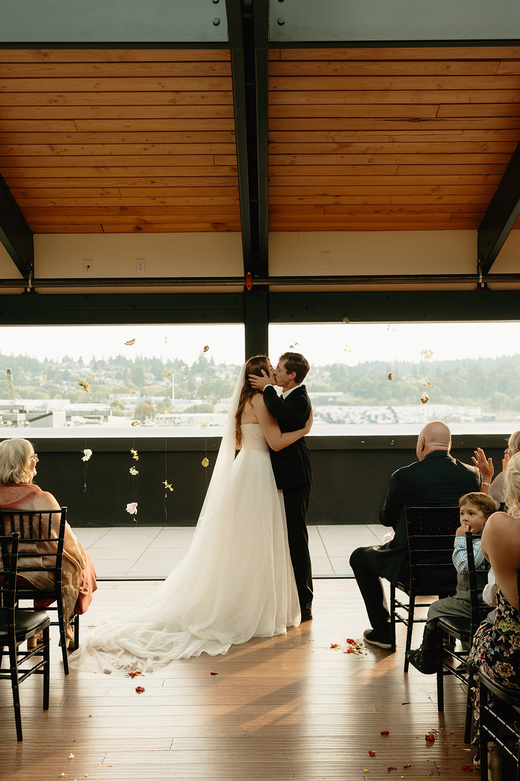 Bride and groom share a first kiss as newlyweds during their wedding in Washington, with flower petals falling around them.