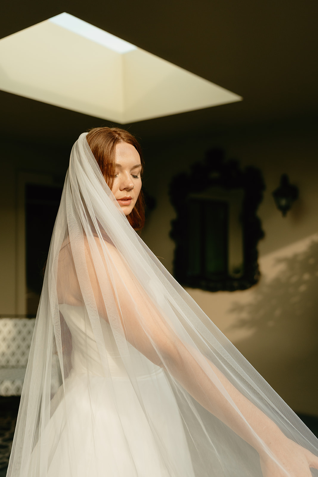 Bride bathed in natural light, standing beneath a skylight with her veil cascading around her.
