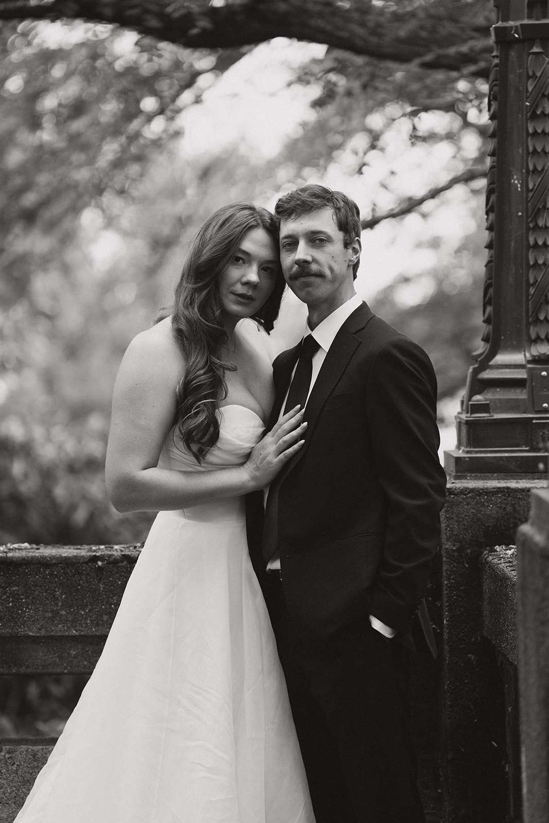Moody black and white portrait of the couple standing together beside an old stone railing surrounded by trees.