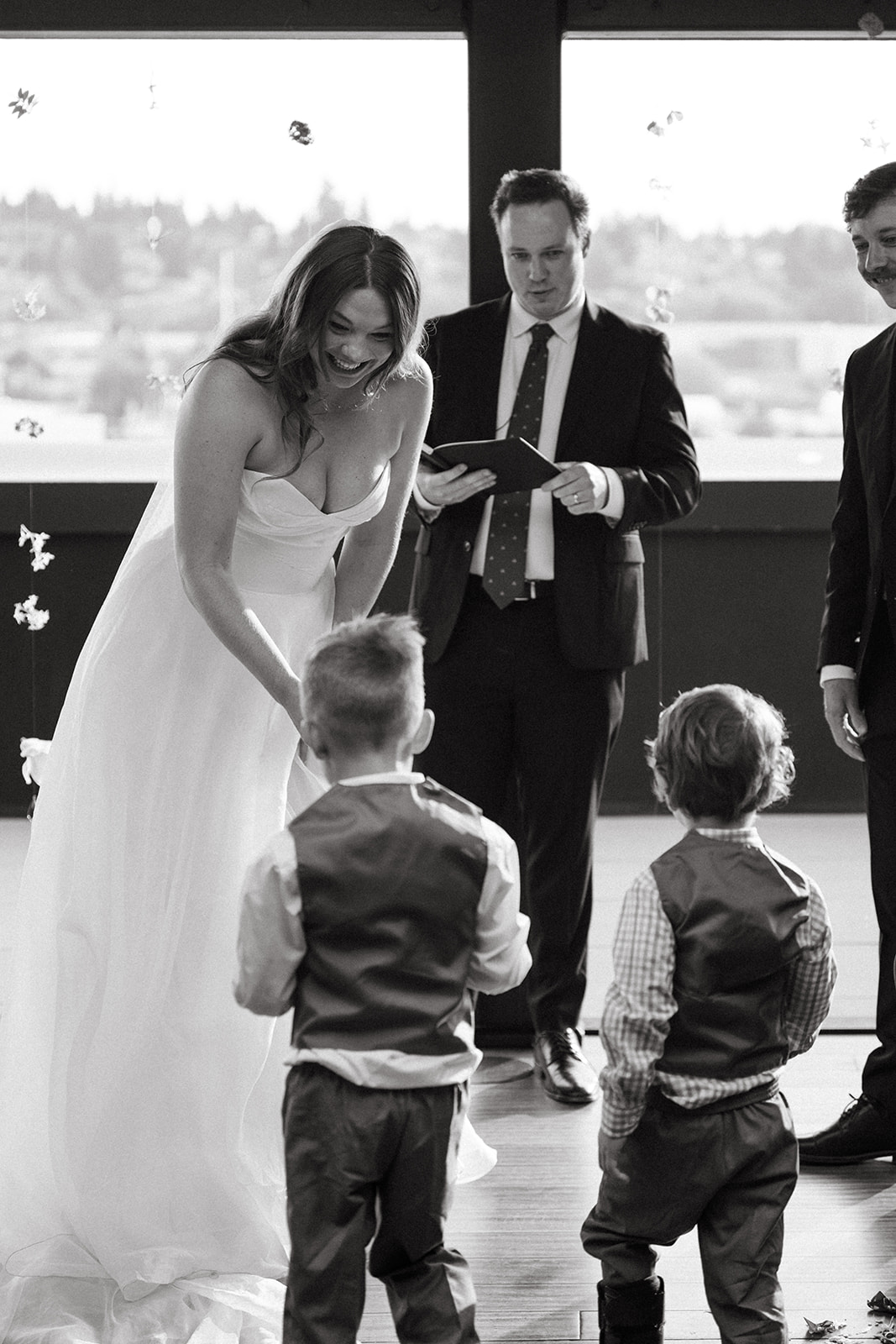 Black and white image of the bride smiling as she bends down to greet two young children at the altar.