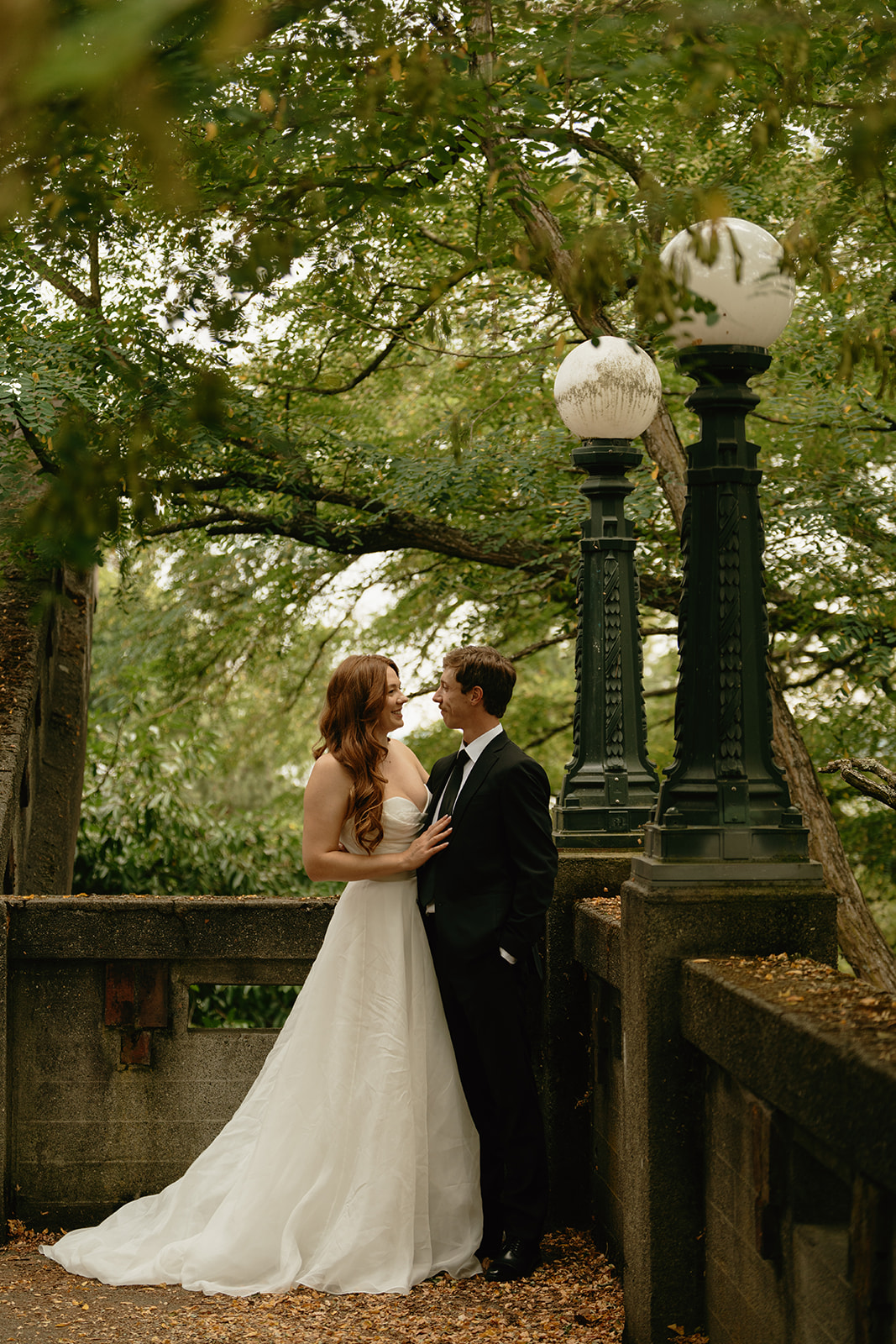 Bride and groom share a quiet moment under vintage lamp posts surrounded by lush greenery in an old garden walkway.