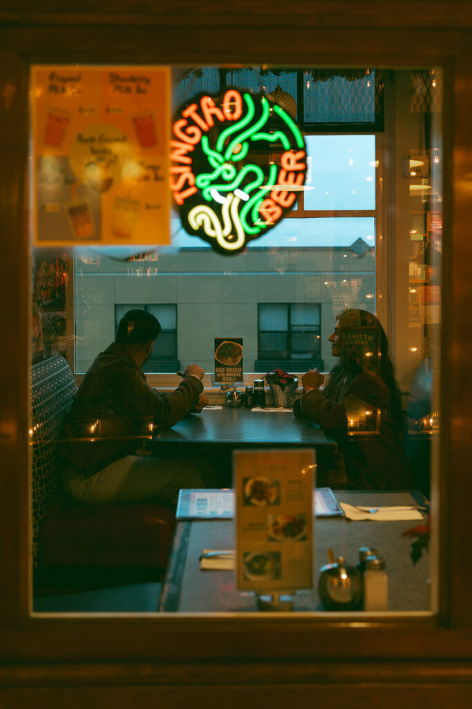 A cinematic couple at a diner in Pike Place Market, perfectly framed in the window and surrounded by bright neon night lights